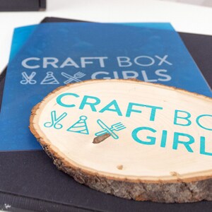 May include: A round wooden sign with the text "CRAFT BOX GIRLS" in teal blue lettering. The sign is decorated with a pair of scissors, a party hat, and a knife and fork.