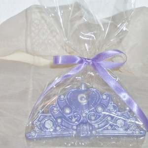 May include: A lavender-colored tiara-shaped soap, embellished with silver accents and a central initial, is encased in clear cellophane. The package is tied with a matching lavender ribbon, creating a gift-like presentation. The soap is designed to resemble a princess crown.