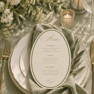 May include: An oval-shaped paper menu with a green border, featuring the text Menu, Starter, Main, and Dessert, placed on a sage green fabric napkin atop a white plate. The menu lists food items including caprese skewers, lamb, and cheesecake.