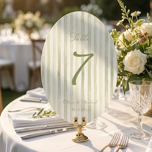 K&ouml;nnte beinhalten: Eine ovale Tischnummerkarte mit einem gr&uuml;n-wei&szlig;en Streifenmuster. Die Karte zeigt die Nummer 7, das Wort "Table" und die Namen Olivia & Michael mit dem Datum 08.10.2027. Die Karte wird von einem goldenen St&auml;nder gehalten.