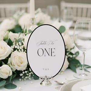 May include: An elegant table setting with a white oval table number card that reads "Table No ONE" in black script. The card is set in a silver stand, surrounded by white roses, greenery, and glassware. A formal event theme.