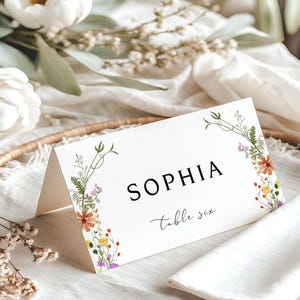 May include: A white place card with the name "SOPHIA" in black, and "table six" in script. The card is decorated with floral illustrations in orange, yellow, and purple. It rests on a woven tray with a white napkin and flowers.