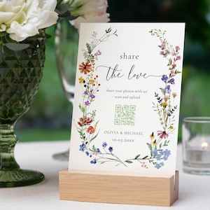 Könnte beinhalten: Hochzeitsempfangsschild mit den Worten "share the love" und floralem Kranzdesign. Das Schild enthält einen QR-Code und die Namen Olivia & Michael mit einem Datum. Das Schild wird auf einem Holzblock angezeigt.