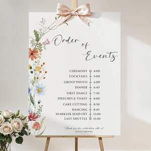 May include: A white wedding sign with a floral border and a list of events with corresponding times. The sign reads "Order of Events" and includes events such as "Ceremony", "Cocktails", "Group Photo", "Dinner", "First Dance", "Speeches & Toast", "Cake Cutting", "Dancing", "Sparkler Send Off", and "Last Shuttle".