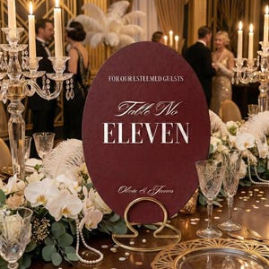 Op de afbeelding: Een bordeauxrode tafelnummerbord met de tekst "Table No. ELEVEN" en "Olivia & James" in witte letters. Het bord wordt vastgehouden door een gouden standaard en is omgeven door bloemstukken en kaarsen.