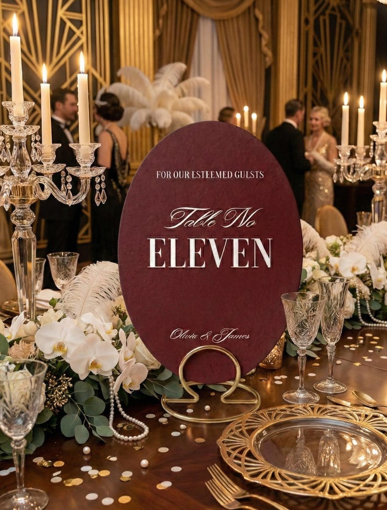 Op de afbeelding: Een bordeauxrode tafelnummerbord met de tekst "Table No. ELEVEN" en "Olivia & James" in witte letters. Het bord wordt vastgehouden door een gouden standaard en is omgeven door bloemstukken en kaarsen.