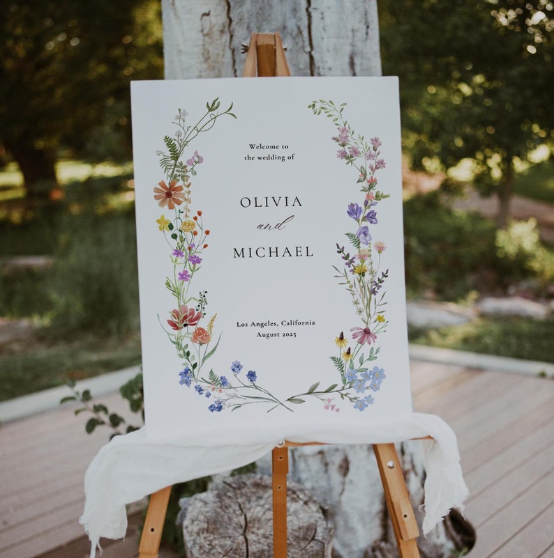May include: A white wedding sign with a floral wreath border. The sign reads "Welcome to the wedding of Olivia and Michael, Los Angeles, California, August 2023."