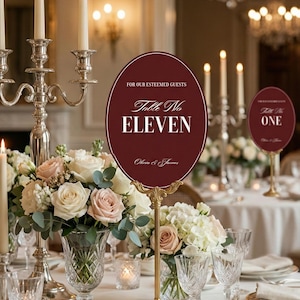 Op de afbeelding: Elegante bruilofttafelsetting met bloemstukken, kaarsen en kristallen glazen. Een bordeauxrode ovale tafelnummerbord met gouden standaard leest "Table No. Eleven" en "For Our Esteemed Guests".