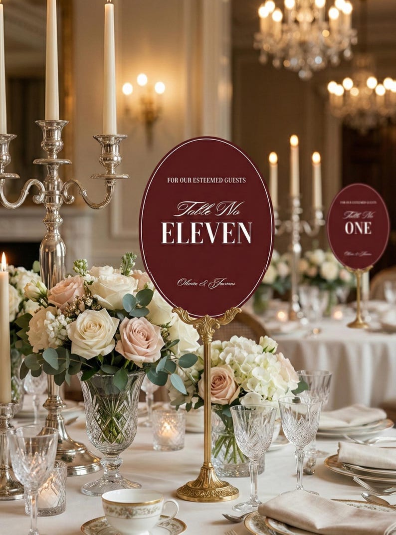 Op de afbeelding: Elegante bruilofttafelsetting met bloemstukken, kaarsen en kristallen glazen. Een bordeauxrode ovale tafelnummerbord met gouden standaard leest "Table No. Eleven" en "For Our Esteemed Guests".