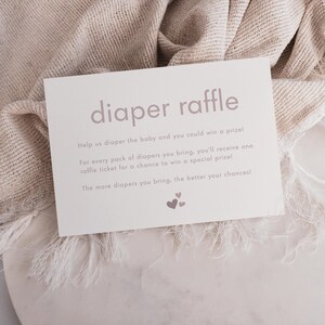 May include: A white card with the text "diaper raffle" in pink lettering. The card has instructions for a diaper raffle, encouraging guests to bring diapers for a chance to win a prize. The text reads: "Help us diaper the baby and you could win a prize! For every pack of diapers you bring, you'll receive one raffle ticket for a chance to win a special prize! The more diapers you bring, the better your chances!"