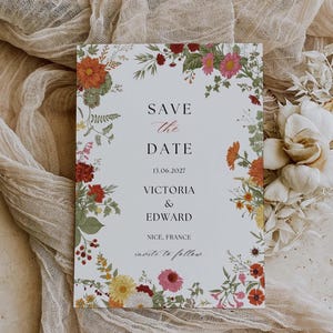 May include: A floral "Save the Date" card with a white background and colorful flowers. The card includes the date, names, and location: "Victoria & Edward, Nice, France". The card is surrounded by a floral border with orange, red, pink, and yellow flowers.