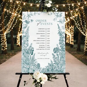 May include: A white sign with a teal floral border and text, displayed on a black easel. The sign reads "Order of Events" and lists the ceremony, cocktails, dinner, and other wedding events. The bottom of the sign says "Thank you for coming!" and the names "Victoria & Edward."