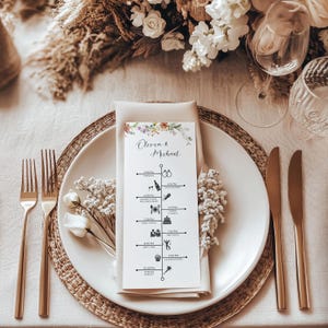 May include: A wedding table setting with a white plate, gold utensils, and a menu card. The menu card features a floral design and a timeline of events. The setting includes a woven placemat, a folded napkin, and decorative flowers.