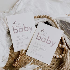 May include: Two white baby shower invitations with a pink and white "oh baby" design. The invitations feature a pink heart design and the text "Amelia Edwards".