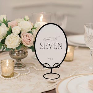 May include: An oval table number card with a black frame and stand, displaying "Table No. SEVEN" in elegant script. The card is set on a table with floral arrangements, candles, and place settings, suitable for a wedding or formal event.