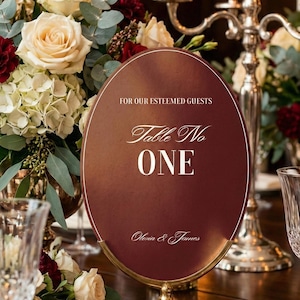 Op de afbeelding: Een bordeauxrode tafelnummerbord met gouden rand en voet, met de tekst "Table No ONE" en de namen "Olivia & James". Het bord staat op een tafel met bloemstukken en kaarsen.