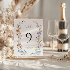 Puede incluir: Una tarjeta de número de mesa de boda floral con marco dorado. La tarjeta presenta una corona de acuarela de flores silvestres coloridas y el texto "mesa 9" y "Olivia y Michael 10.01.2024".