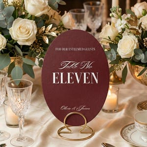 Op de afbeelding: Een bordeauxrode ovale tafelnumerkaart met gouden letters en een gouden metalen standaard. De kaart leest "Table No ELEVEN" en "Voor onze gewaardeerde gasten". De tafelsetting omvat bloemen, kaarsen en glaswerk.