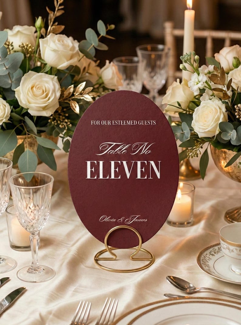 Op de afbeelding: Een bordeauxrode ovale tafelnumerkaart met gouden letters en een gouden metalen standaard. De kaart leest "Table No ELEVEN" en "Voor onze gewaardeerde gasten". De tafelsetting omvat bloemen, kaarsen en glaswerk.