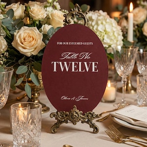 Op de afbeelding: Een bordeauxrode tafelnummerkaart met de tekst "Table No TWELVE" in witte letters, weergegeven op een sierlijke gouden standaard. De kaart luidt "For Our Esteemed Guests" en "Olivia & James". De setting omvat bloemstukken en glaswerk.