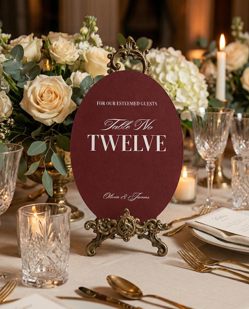 Op de afbeelding: Een bordeauxrode tafelnummerkaart met de tekst "Table No TWELVE" in witte letters, weergegeven op een sierlijke gouden standaard. De kaart luidt "For Our Esteemed Guests" en "Olivia & James". De setting omvat bloemstukken en glaswerk.