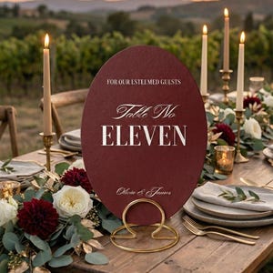 Op de afbeelding: Een bordeauxrode tafelnummerbord met gouden letters met de tekst "Table No ELEVEN" en "Olivia & James". Het bord wordt vastgehouden door een gouden metalen houder en staat op een houten tafel versierd met bloemen en kaarsen.