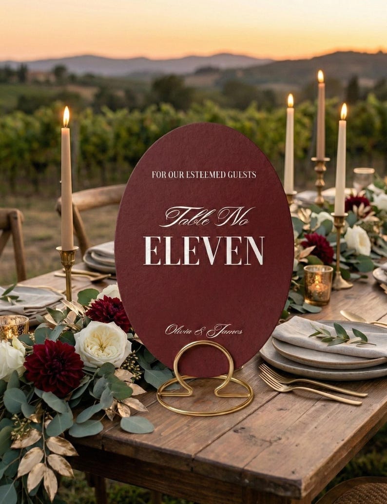 Op de afbeelding: Een bordeauxrode tafelnummerbord met gouden letters met de tekst "Table No ELEVEN" en "Olivia & James". Het bord wordt vastgehouden door een gouden metalen houder en staat op een houten tafel versierd met bloemen en kaarsen.
