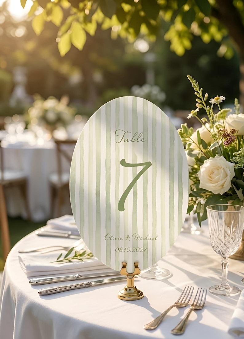 K&ouml;nnte beinhalten: Eine ovale Tischnummerkarte mit einem gr&uuml;n-wei&szlig;en Streifenmuster. Die Karte zeigt die Nummer 7, das Wort "Table" und die Namen Olivia & Michael mit dem Datum 08.10.2027. Die Karte wird von einem goldenen St&auml;nder gehalten.