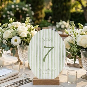 K&ouml;nnte beinhalten: Eine Tischnummernkarte mit gr&uuml;n-wei&szlig;em Streifendesign, mit der Nummer 7. Die Karte steht auf einem Holzsockel, mit den Namen Olivia & Michael und dem Datum 08.10.2027. Der Tisch ist mit Besteck, Gl&auml;sern und Blumen gedeckt.
