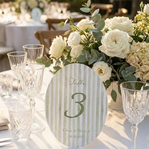 K&ouml;nnte beinhalten: Eine Hochzeitstafel mit Blumenschmuck und Tischnummer. Die ovale Tischnummer hat ein gr&uuml;n-wei&szlig; gestreiftes Design mit der Nummer "3", den Namen "Olivia & Michael" und dem Datum "08.10.2027". Kristallgl&auml;ser und Besteck liegen auf der wei&szlig;en Tischdecke.