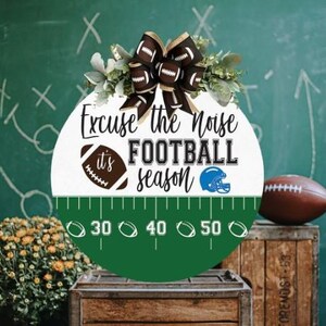 Puede incluir: Un colgador de puerta redondo de madera con un diseño de campo de fútbol verde y blanco. El texto "Excuse the Noise it's FOOTBALL season" está escrito en negro sobre fondo blanco. También se incluye en el diseño un balón de fútbol marrón y un casco de fútbol azul.