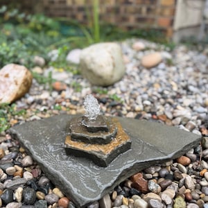 May include: A tiered stone water fountain featuring a flat, dark gray base topped with two smaller, stacked hexagonal stone layers. A small, clear stream of water bubbles from the center of the top tier, creating a wet, glistening surface.