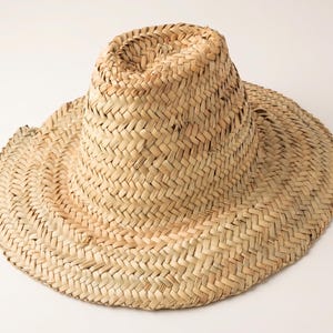 Pode incluir: Um chapéu de palha castanho-claro com textura entrançada, aba larga e plana e copa alta e arredondada. O chapéu é feito de fibras vegetais secas naturais dispostas num padrão de espinha de peixe consistente e entrelaçado em toda a superfície.