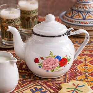 Puede incluir: Tetera de esmalte blanco con un diseño floral con rosas rosas, rojas y azules. La tetera tiene un borde y un asa negros, y está colocada sobre un mantel estampado colorido. Otros elementos incluyen vasos y una jarra blanca.