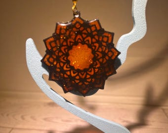 Könnte beinhalten: Ein hängender Ohrring mit einem kreisförmigen, sternförmigen Anhänger, der ein komplexes, dunkelbraunes Mandala-Muster über einem leuchtend orangefarbenen, glitzernden Zentrum aufweist und an einem goldfarbenen Metallhaken befestigt ist.