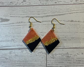 Könnte beinhalten: Ein Paar rautenförmige Ohrhänger mit einem farblich abgesetzten Design, bestehend aus einem korallenfarbenen oberen Teil und einem schwarzen unteren Teil, die durch ein horizontales Band aus goldenem Glitzer getrennt sind, an Goldhaken.