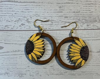 Könnte beinhalten: Ein Paar kreisförmige Holzohrringe mit Sonnenblumenmotiv. Jeder Ohrring besteht aus einem dunkelbraunen Holzringrahmen mit einem aufgedruckten gelb-braunen Sonnenblumenblütenblatt-Design am Innenrand sowie goldfarbenen Metallhaken.