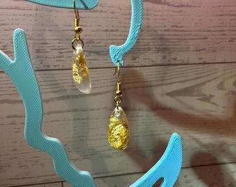 Könnte beinhalten: Ein Paar Ohrhänger mit tropfenförmigen Elementen aus klarem Harz, die mit Blattgoldflocken durchsetzt sind. Die Ohrringe sind an goldfarbenen Metallhaken befestigt und zeichnen sich durch ein durchscheinendes Design mit goldenen Akzenten aus.