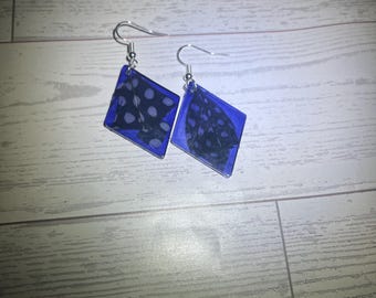 Könnte beinhalten: Ein Paar rautenförmige Ohrringe mit einem Rahmen aus durchscheinendem blauem Harz. Jeder Ohrring enthält einen dunklen Stoffeinsatz mit weißem Punktmuster auf der einen und einem dunklen, strukturierten Blumenmuster auf der anderen Seite, mit Silberhaken.