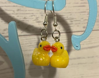 Könnte beinhalten: Ein Paar Ohrhänger mit kleinen, leuchtend gelben Quietscheentchen-Anhängern mit orangefarbenen Schnäbeln und schwarzen Augen. Die Enten hängen an silberfarbenen Metallhaken und bilden ein verspieltes und auffälliges Accessoire für die Ohren.