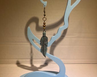 Könnte beinhalten: Ein einzelner Ohrhänger mit einem bronzefarbenen Metallhaken und kleinen runden Perlen, verbunden mit einem strukturierten, oxidierten türkis-grünen Anhänger in Federform, der an einem hellblauen, geschwungenen Ständer hängt.