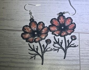 Könnte beinhalten: Ein Paar Hängeohrringe mit einer schwarzen Blumensilhouette, deren Blütenblätter ein schimmerndes, glitzerndes korallenrosa Zentrum aufweisen. Das Design zeigt eine Blume an einem Stiel mit einer kleinen Knospe, befestigt an silberfarbenen Ohrhaken.