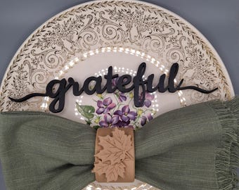 Könnte beinhalten: Ein weißer Keramikteller mit goldenem Blumenmuster zeigt den Schriftzug grateful als schwarzes Ausschnitt-Element. Eine salbeigrüne Leinen-Serviette mit Fransen wird von einem hellbraunen Serviettenring mit zwei Ahornblatt-Motiven gehalten.