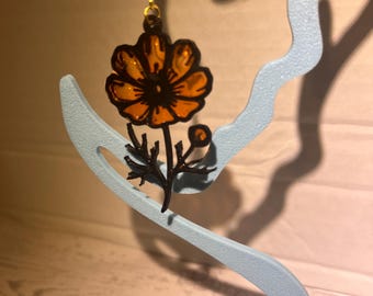 Könnte beinhalten: Ein hängender Ohrring mit einer durchscheinenden orangefarbenen Blume mit schwarzen Umrissen und einem schwarzen Stiel. Das florale Design hängt an einem goldfarbenen Metallhaken. Das Stück zeigt einen leuchtenden Orangeton in den Blütenblättern, der durch kräftige schwarze Details kontrastiert wird, die die Form der Blume, des Stiels und der Blätter definieren.