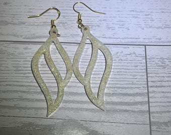 Könnte beinhalten: Ein Paar Ohrhänger aus durchscheinendem, weißem Harz in Tropfenform mit offener Mitte und eingebettetem Goldglitter. Die Ohrringe sind an goldfarbenen Metallhaken für durchstochene Ohrläppchen befestigt und erzeugen ein helles, reflektierendes Aussehen.