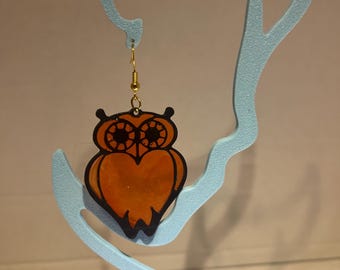 Könnte beinhalten: Ein eulen-förmiger Ohrring mit einem durchscheinenden orangefarbenen Körper und einer schwarzen Umrandung. Das Design zeigt detaillierte schwarze kreisförmige Augen und eine spitze Ohrensilhouette. Der Ohrring ist an einem goldfarbenen Haken befestigt.