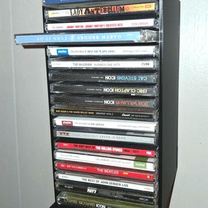 May include: A black wall-mounted CD storage rack holding a vertical stack of music albums. One CD case is partially pulled out from the shelf. The collection includes titles by Garth Brooks, Lady Antebellum, Johnny Horton, and The Beatles.