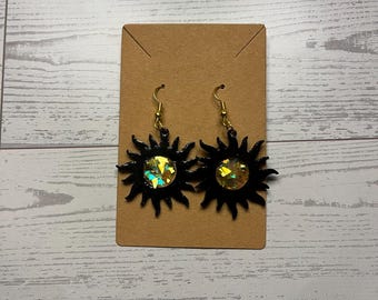Könnte beinhalten: Ein Paar schwarze, sonnenförmige Ohrringe mit goldfarbenen Metallhaken und einer zentralen, kreisförmigen Einlage, die mit irisierenden, mehrfarbigen Folienflocken gefüllt ist, die das Licht einfangen, auf einer braunen Pappkarte.