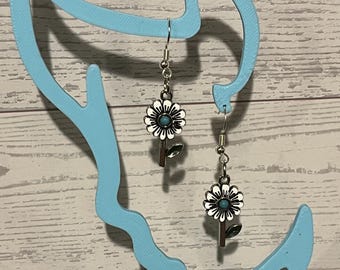 Könnte beinhalten: Ein Paar silberfarbene Ohrhänger mit einem weißen Blumenanhänger, einer zentralen türkisfarbenen Perle und einem einzelnen grünen Blatt am Stiel. Die Ohrringe hängen an silberfarbenen Haken und werden auf einem hellblauen Ständer präsentiert.