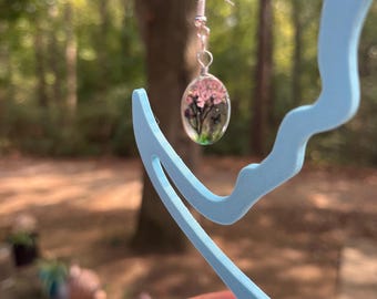 Könnte beinhalten: Ein ovaler Ohrring aus Kunstharz mit einem Miniatur-Landschaftsdesign, in dem rosa Blüten und grünes Blattwerk eingeschlossen sind. Der Ohrring hängt an einem silberfarbenen Metallhaken und wird auf einem hellblauen, schwanenförmigen Ständer präsentiert.